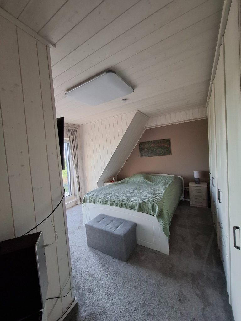Schlafzimmer mit viel Tageslicht – Haus Oederquart Schlafzimmer mit einem Bett, einem Nachttisch und einem grauen Hocker. Helle Wände und Fenster.